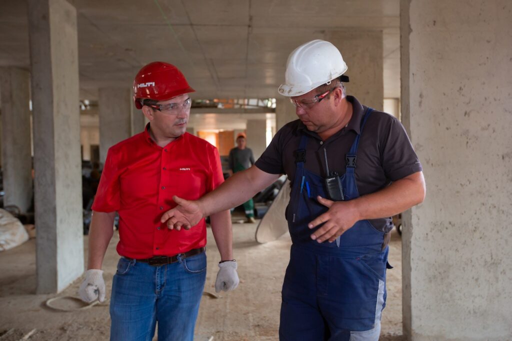 Two men in hardhats walk through construction area having conversation