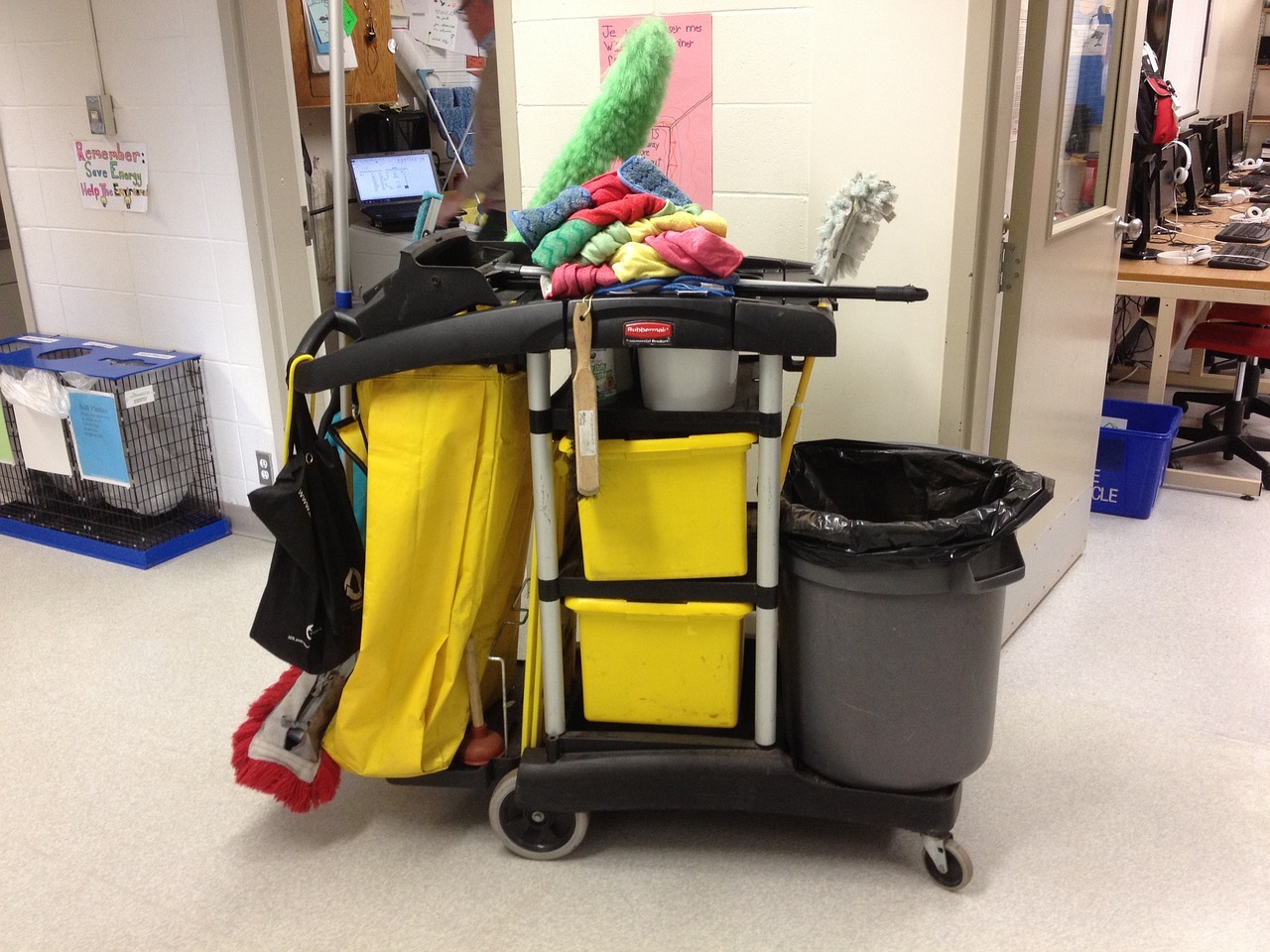Janitorial cart with towels on top and a large garbage bin on the right. The cart is parked in front of a door of a room full of desks and computers