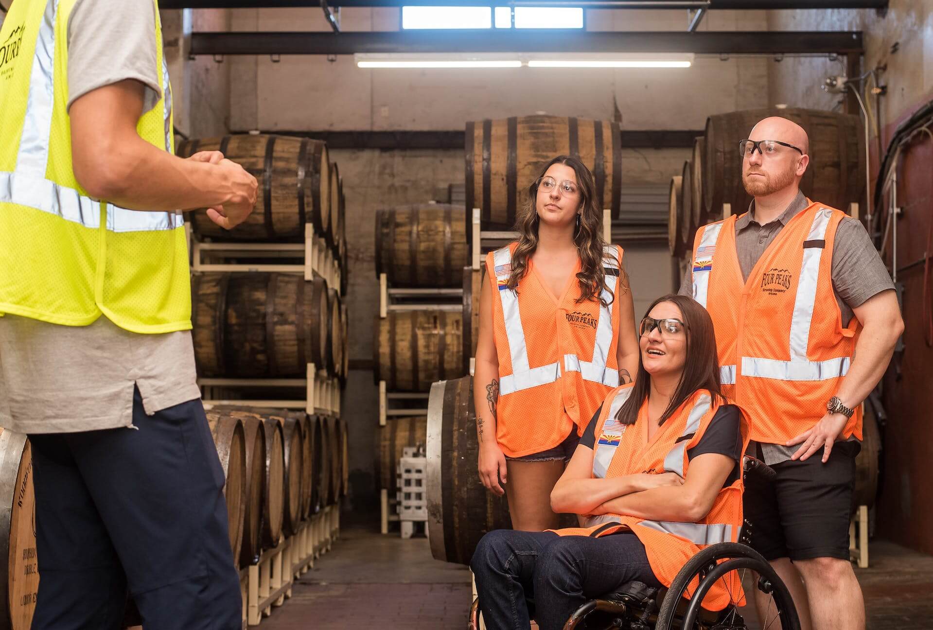 Manager in yellow vest in foreground gives direction to 3 employees in orange vests in the background. One of the employees is in a wheelchair